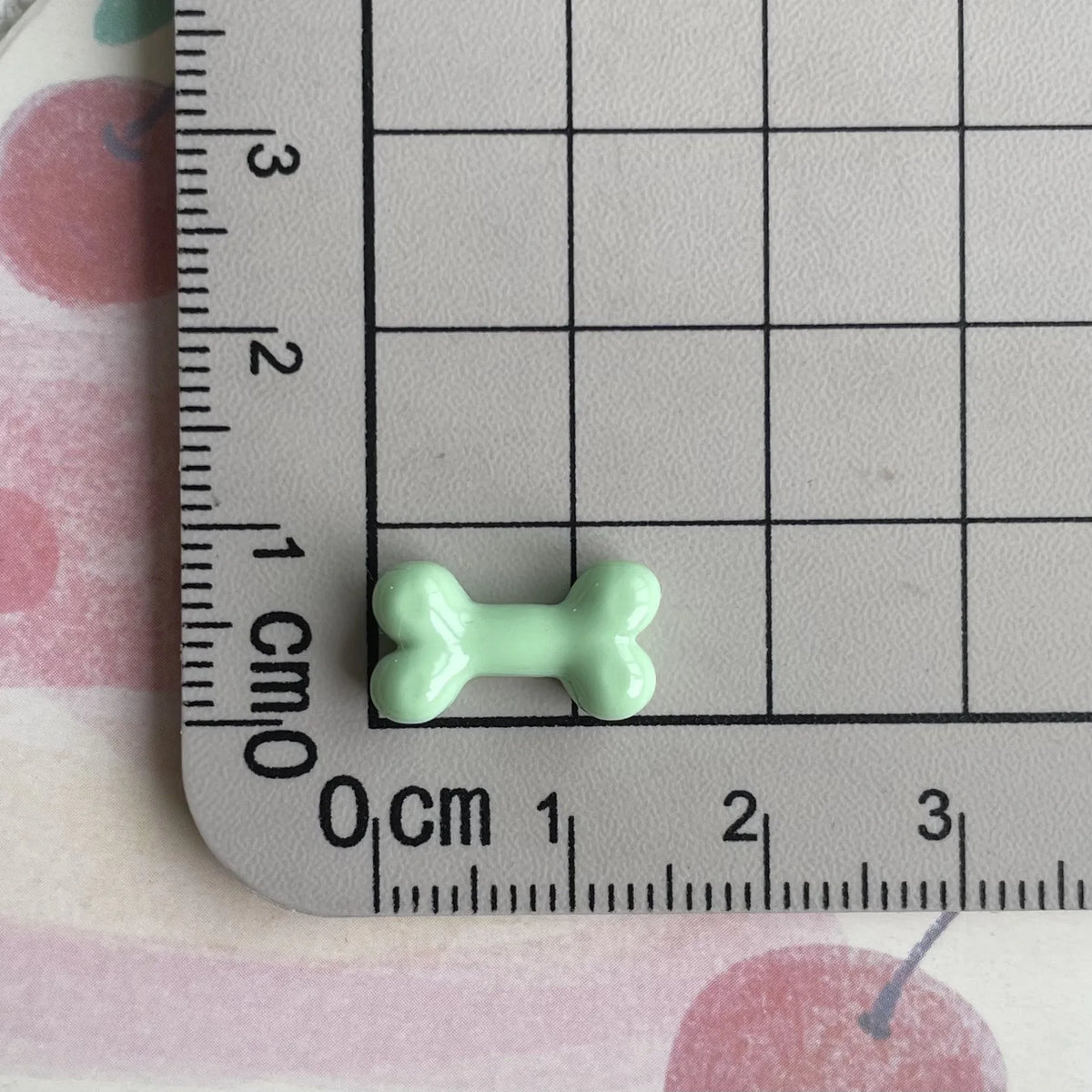 A small, light green resin bone charm is placed on a ruler marked in centimeters.