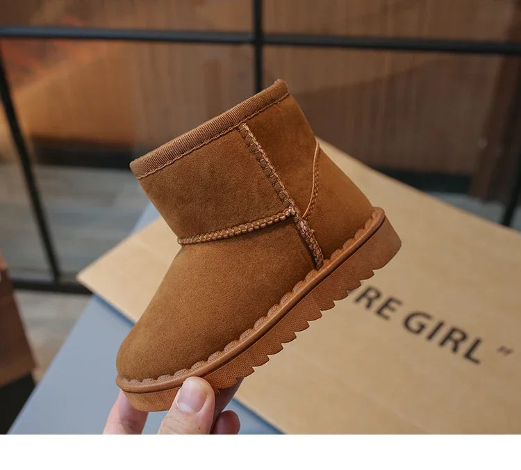 A close-up of a tan suede slip-on winter boot for a child, held against a brown cardboard box.