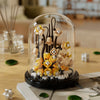 A glass dome display of a buildable eternal rose flower kit featuring small yellow and white flowers on a black base.