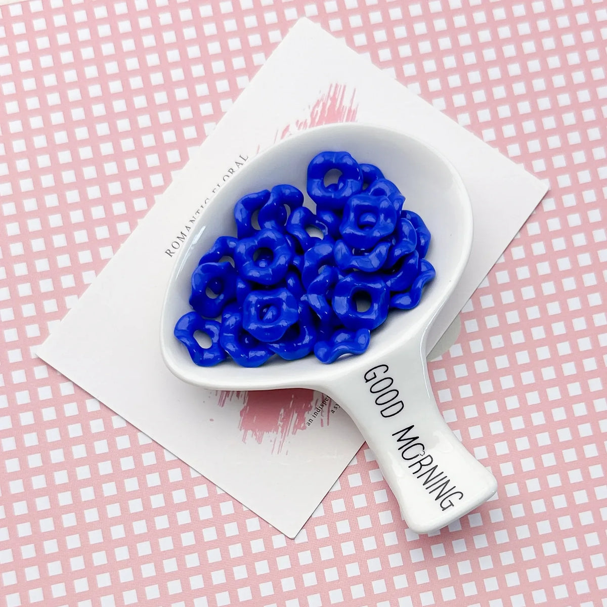 A white ceramic spoon holds a cluster of bright blue acrylic beads with a floral edge on a pink patterned surface.