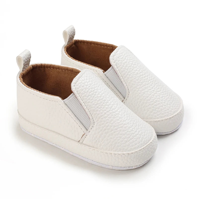 A pair of white baby first walker sneakers with a dinosaur print detail and brown lining.