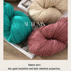 Three skeins of alpaca yarn in teal, beige, and dusty rose colors are displayed on a wooden surface.