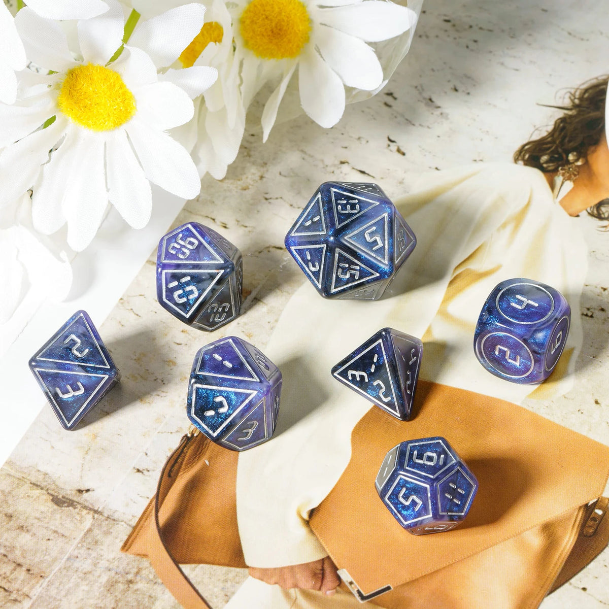 A set of seven blue resin polyhedral dice displayed on a light surface next to a white cloth and a brown leather folder.