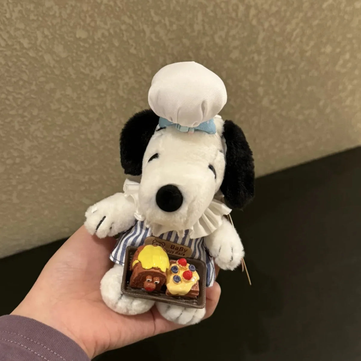 A white Snoopy plush toy with black ears and a blue bow is held in a hand, featuring a small charm.