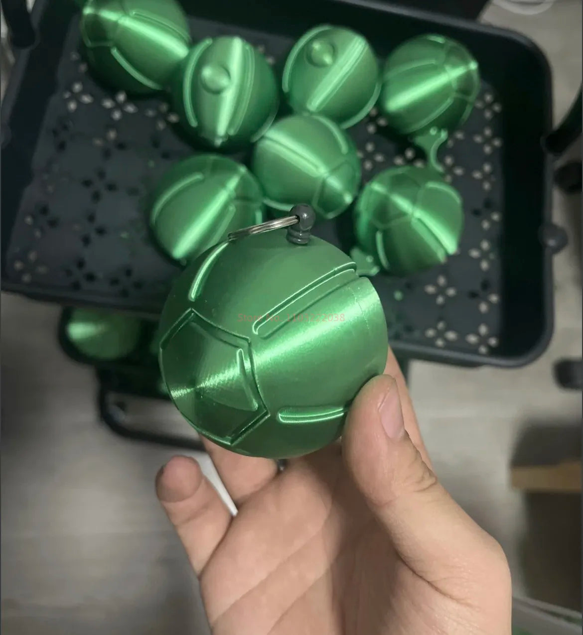 A hand holds a green 3D printed steel ball fidget toy, surrounded by other similar balls on a dark tray.