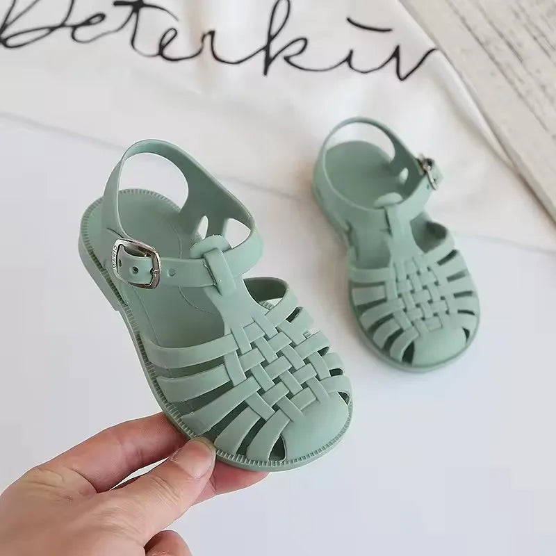 A hand holds a pair of mint green girls' gladiator jelly sandals with woven straps and non-slip soles.