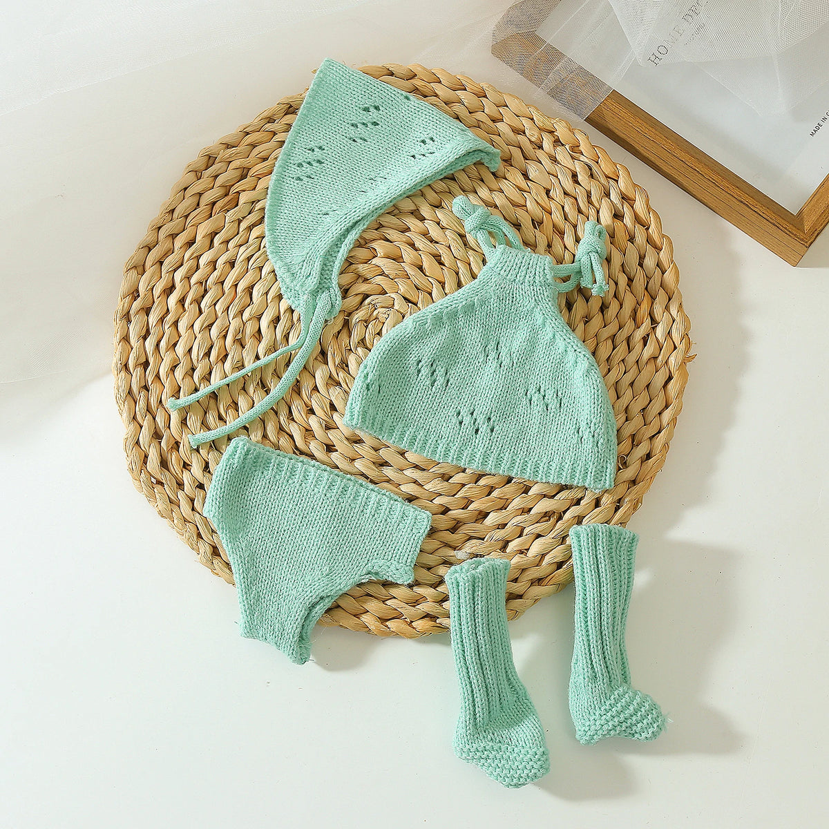 A set of three light mint green knitted doll outfits including a hat, dress, and boots displayed on a woven mat.