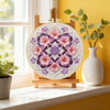A wooden display holds a circular embroidery piece featuring pink and purple floral designs on a light background.