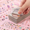 A hand is holding a stack of 12 landscape stickers on a pink background.