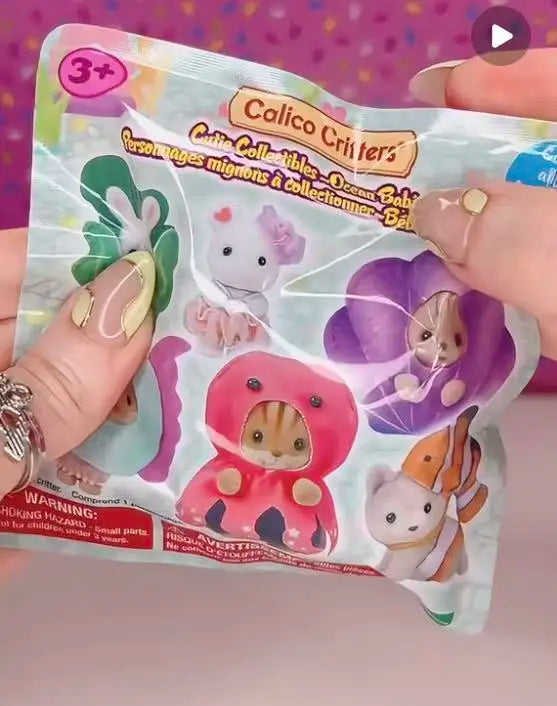 A hand holds a package of Calico Critters figures, including a pink and purple animal figure.