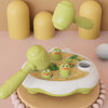 A green and yellow Montessori animal hammer toy with various plastic parts is shown on a light wooden surface.