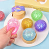 A hand holds a pink glitter squishy bun next to various colored glitter balls and a wooden mold.