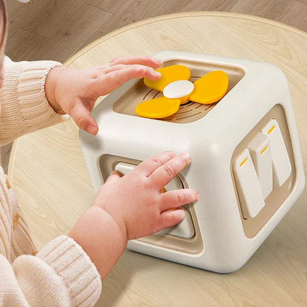 A toddler is interacting with a white Montessori activity cube featuring yellow and white manipulative pieces on a wooden sur