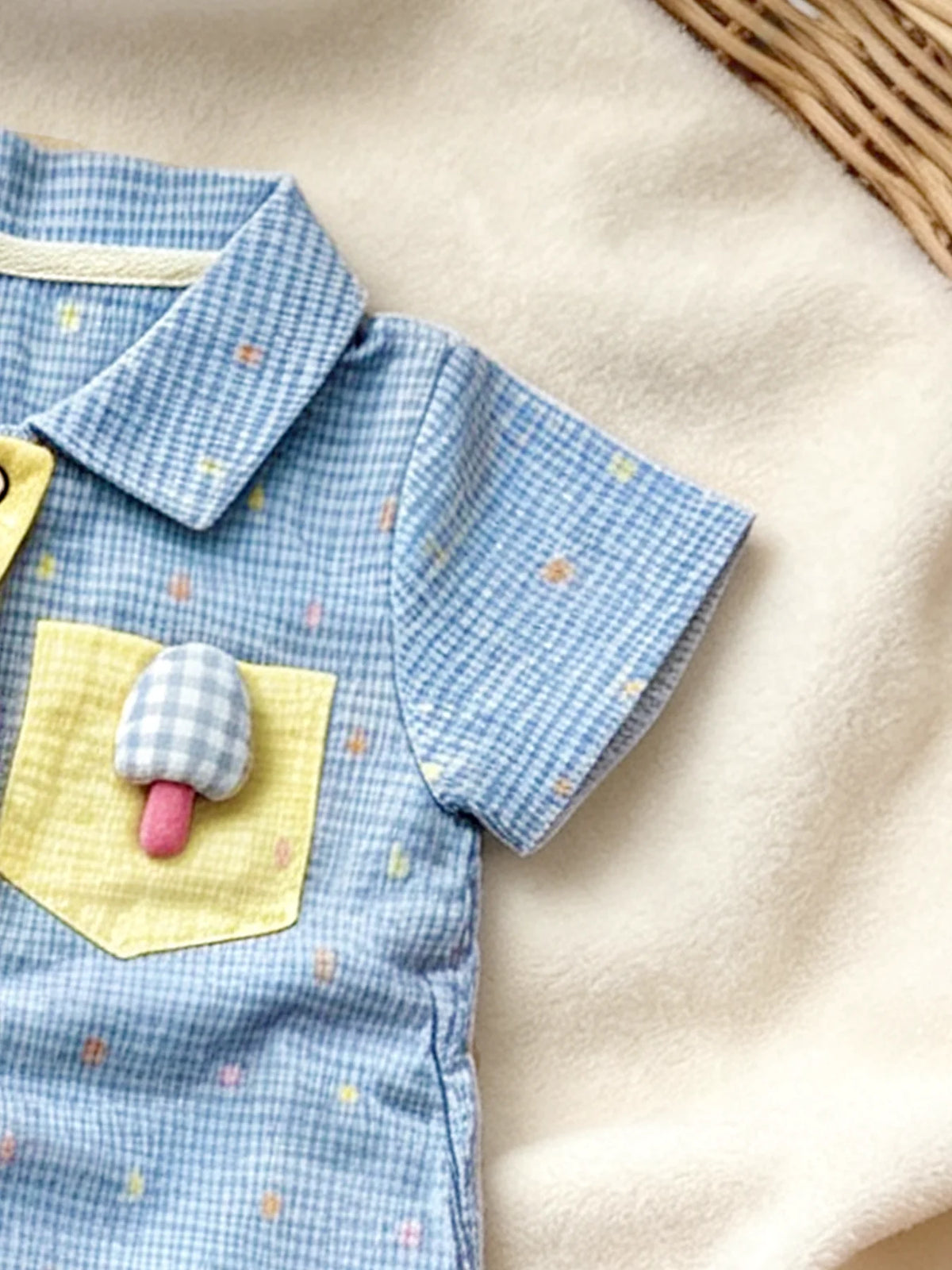 A baby boy short-sleeve romper in blue gingham with yellow appliqués of a bear and mushroom on a cream fabric background.