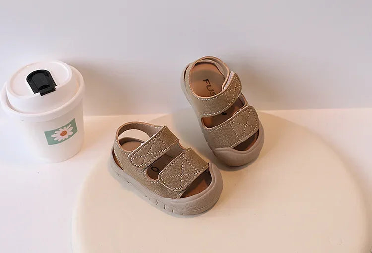 A pair of beige suede toddler sandals for boys and girls resting next to a white cup.