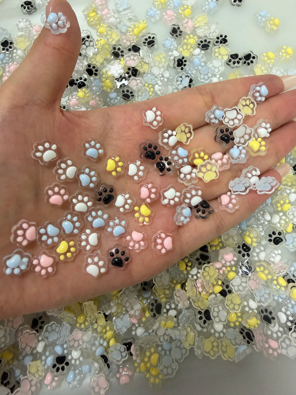 A hand displays a collection of luminous cat paw nail art charms in various pastel colors for DIY crafts.
