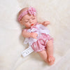 A baby doll wearing a pink ruffled jumpsuit outfit with a matching bow and headband, lying on a white fluffy surface.
