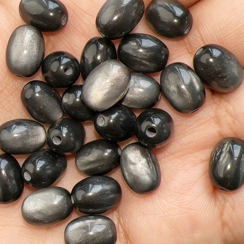 A close-up of ten dark gray and black resin starry sky cat's eye beads resting on a person's hand.
