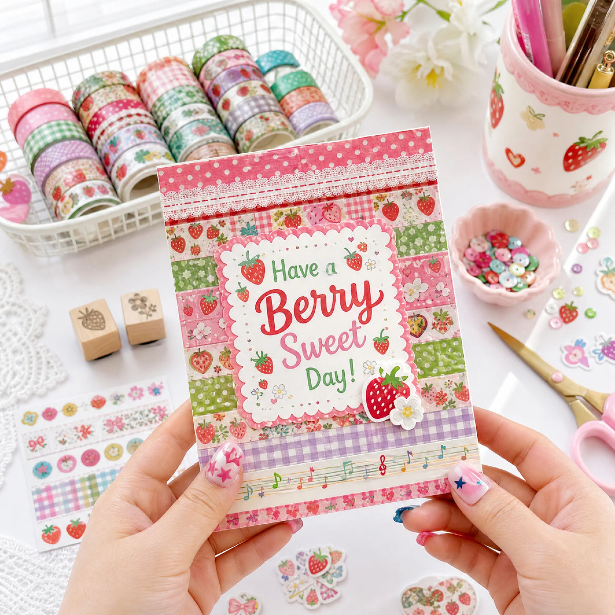 A hand holds a pink and green patterned Washi tape set with a strawberry design, next to a white basket of craft supplies.