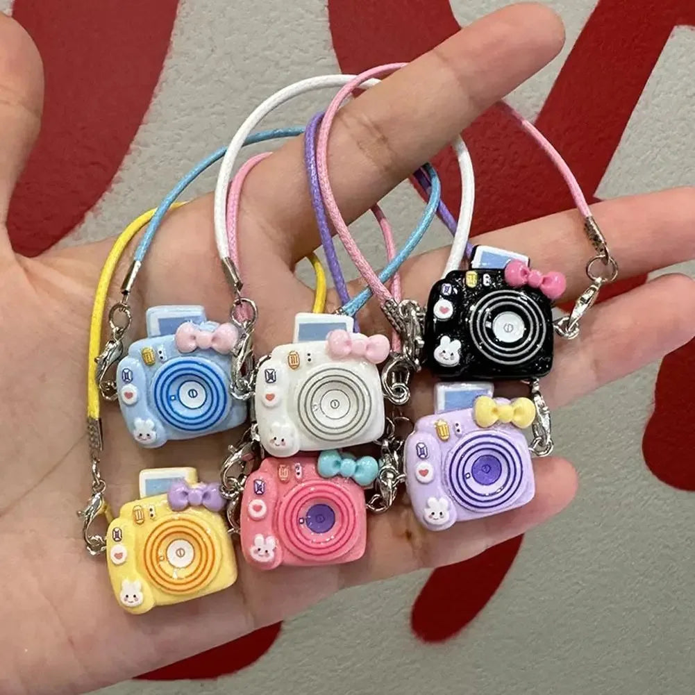 A hand holds a necklace with various colorful mini plush camera doll accessories on pink and white cords.
