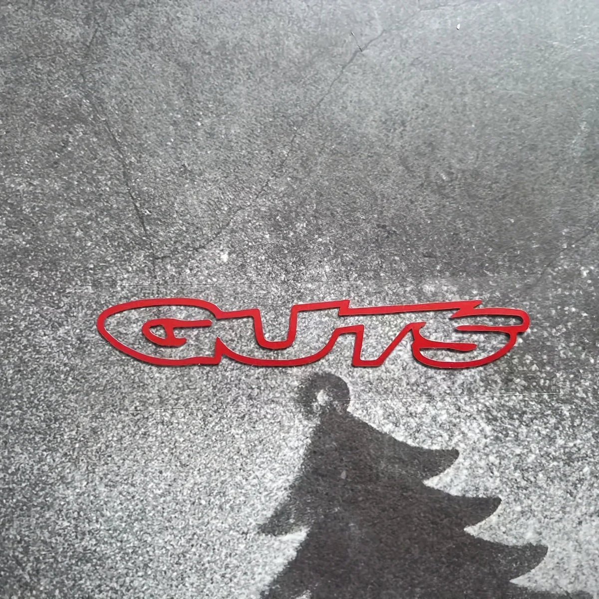 A red silhouette of the word GUTS is reflected on a wet, reflective surface.