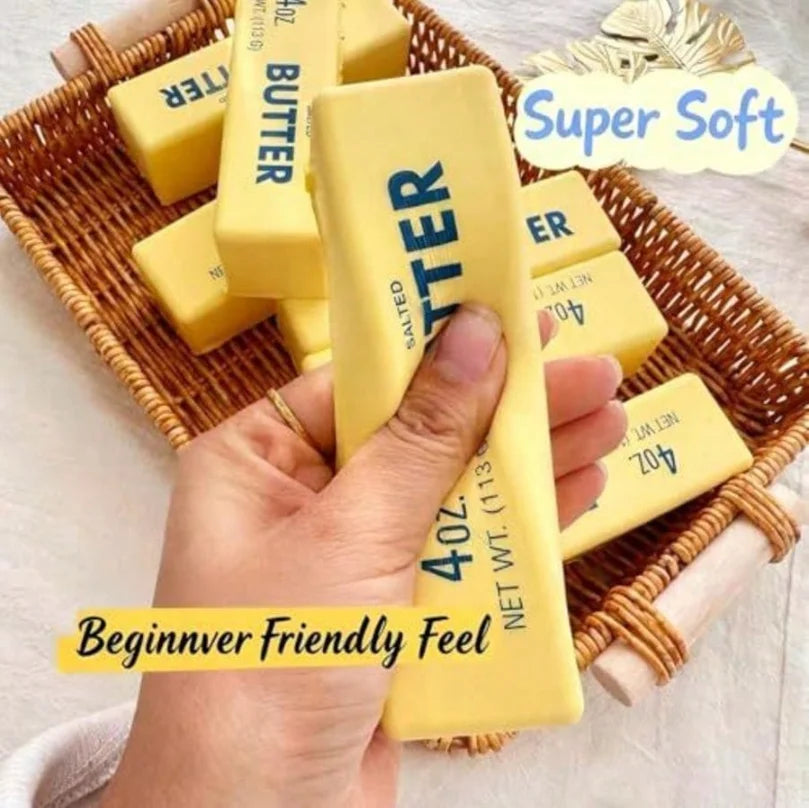 A hand holds a yellow butter stick squishy toy in a woven basket with a Super Soft label.