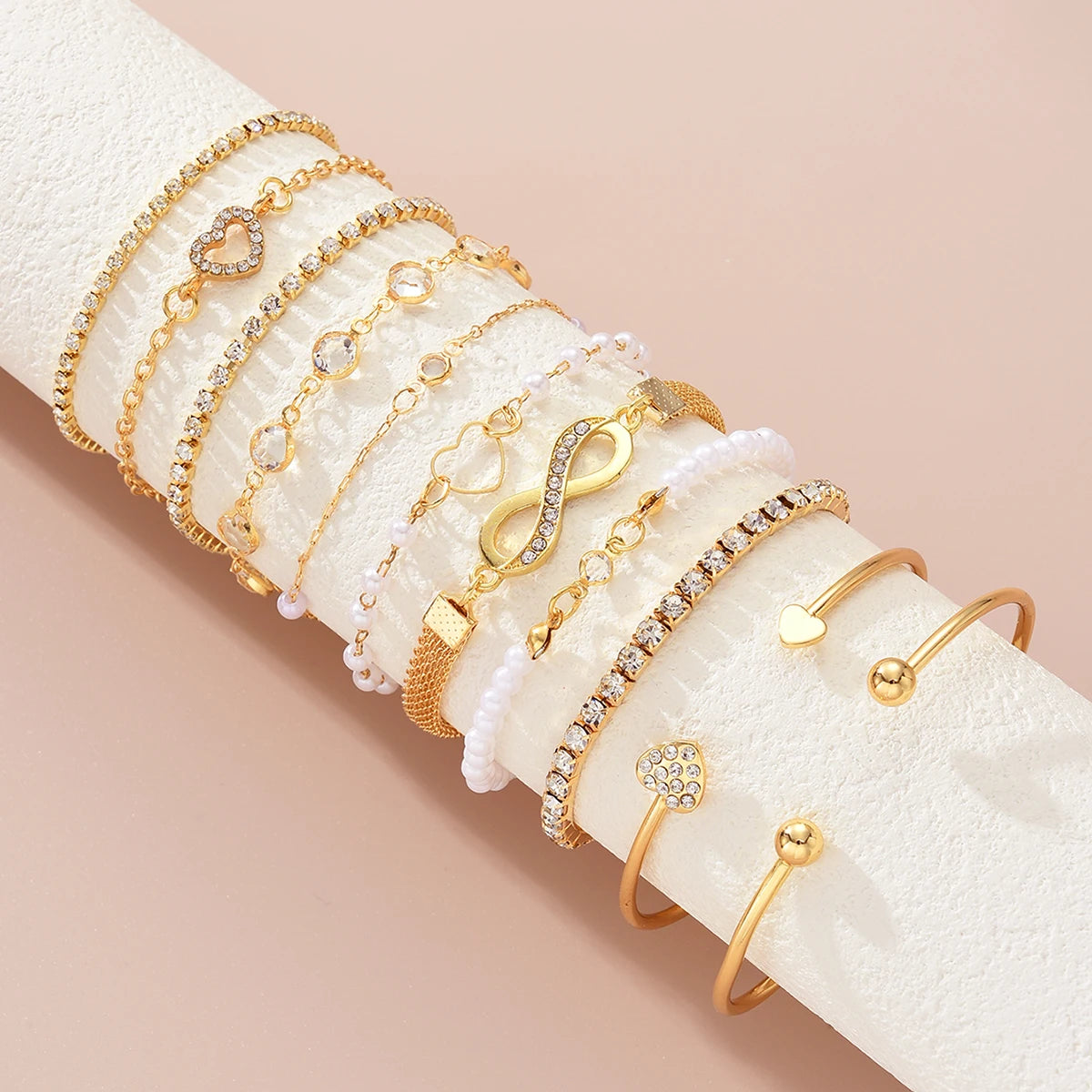 A collection of 11 gold and crystal bracelets featuring heart and infinity signs stacked on a white wrist.