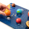 A hand places an orange sphere onto a blue wooden solar system puzzle board showing planets like Mercury, Earth, Venus, and M