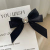 A black satin bow with a clear rhinestone heart embellishment displayed on a white card.