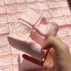 A hand is squeezing a pink strawberry milk squishy stress relief toy on a pink grid surface.