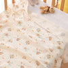A light beige baby gauze swaddle blanket with a pattern of small bears and rainbows, displayed next to soft stuffed animals.