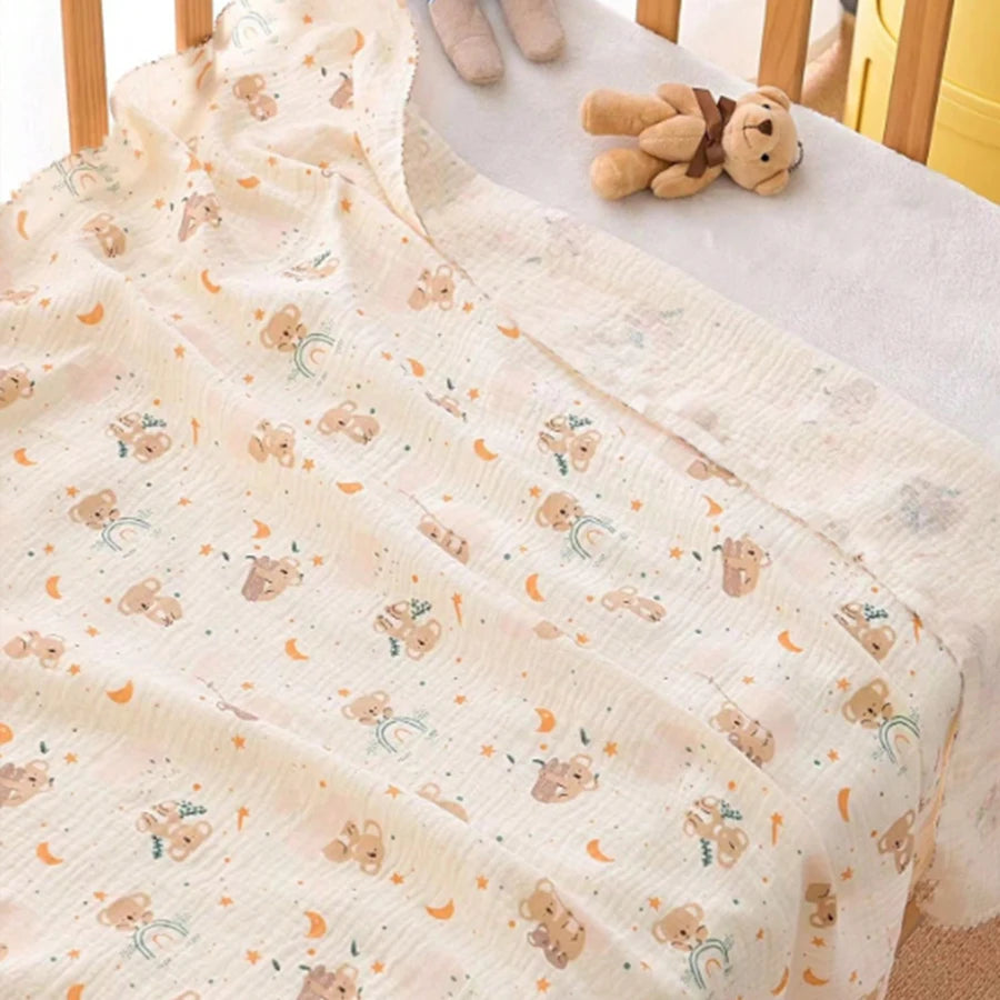 A light beige baby gauze swaddle blanket with a pattern of small bears and rainbows, displayed next to soft stuffed animals.