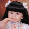 A close-up of a lifelike reborn baby doll with soft cloth body featuring black hair and white floral accessories.