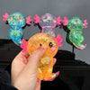 A hand holds an orange squishy axolotl stress relief toy with pink accents against a dark background.