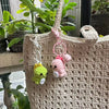 A beige crochet dinosaur keychain hangs from a macrame cord on a woven fabric bag.