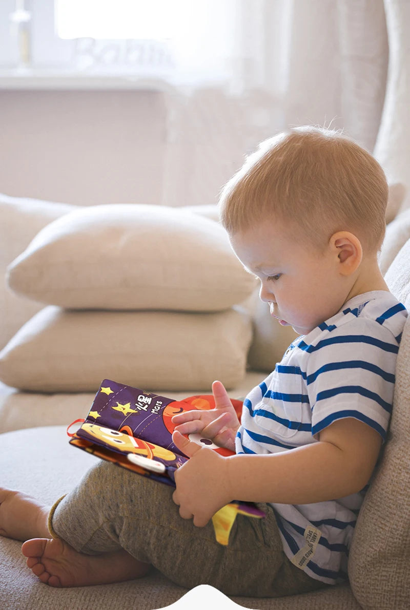 A toddler sits on a couch looking at a soft sensory cloth book for babies with crinkle pages.
