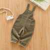 A pair of olive green baby cotton overalls with adjustable straps lying on a wooden surface next to a camera.
