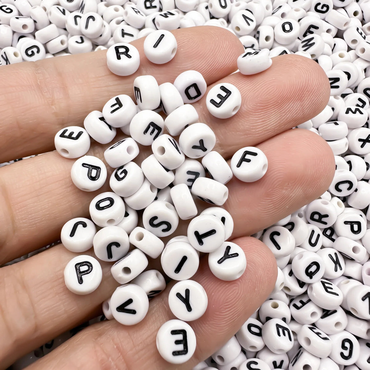 A hand holds a cluster of white acrylic alphabet letter beads for jewelry making on a bed of similar beads.