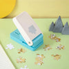 A white puzzle making tool rests on a teal base, surrounded by small gold and white puzzle pieces on a light green surface.