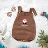 A brown knit baby romper embroidered with a Santa face is displayed on white fur, with matching shoes nearby.