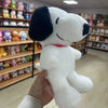 A hand is holding a white and black Snoopy cartoon plush toy with a red collar in a retail store setting.
