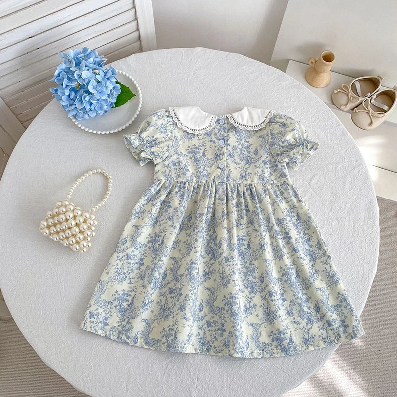 A light blue floral bubble sleeve dress for girls displayed on a white table with pearls and a wooden cup.