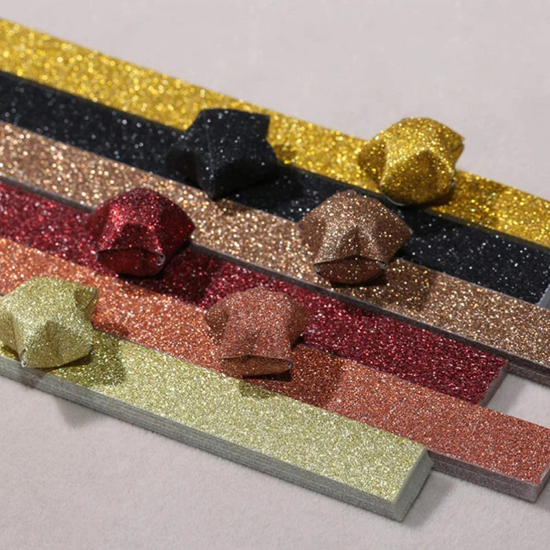 A collection of glitter origami star paper strips in various colors like gold, black, silver, and rose gold arranged diagonal