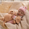 A realistic baby doll wearing a pink knitted hat and a pink romper, cuddling a fluffy brown stuffed animal.