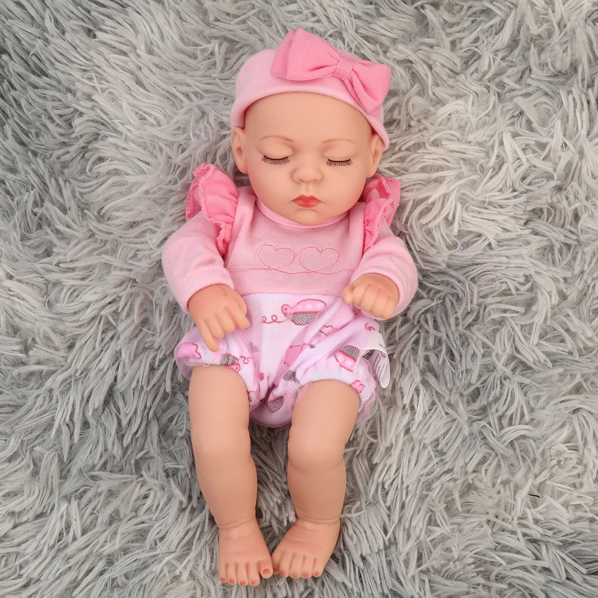 A lifelike soft vinyl reborn doll with a pink outfit and matching pink bow lying on a gray fuzzy surface.