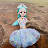 A small mermaid doll with teal hair and a sparkly light blue dress is placed on a brown rock.