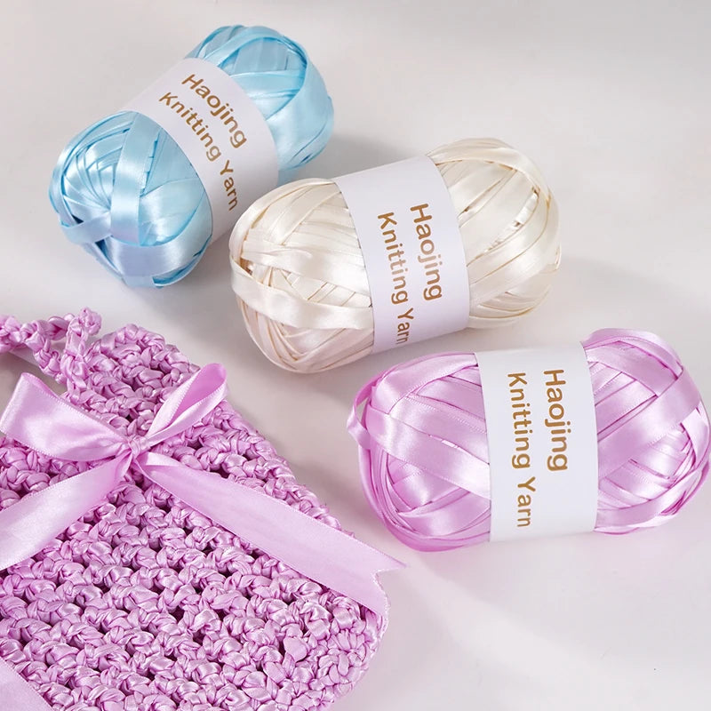 A set of three spools of various colored satin ribbon yarn for knitting and weaving displayed on a white surface.