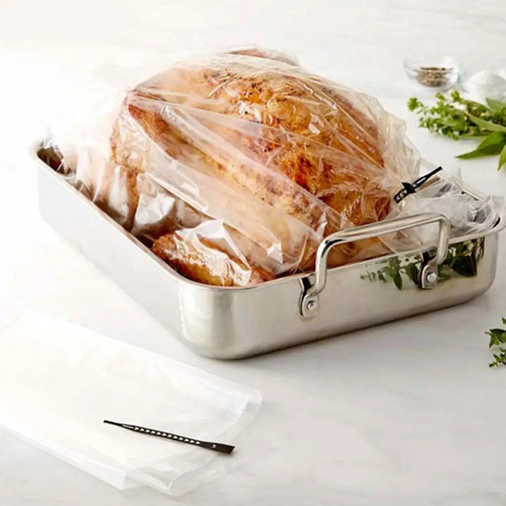 A silver roasting pan holds a turkey or chicken sealed in a heat-resistant oven bag on a white surface.