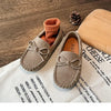 A pair of taupe girls' moccasins with bow details resting on a white cloth next to a wooden board.