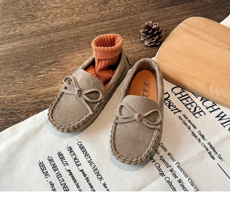 A pair of taupe girls' moccasins with bow details resting on a white cloth next to a wooden board.